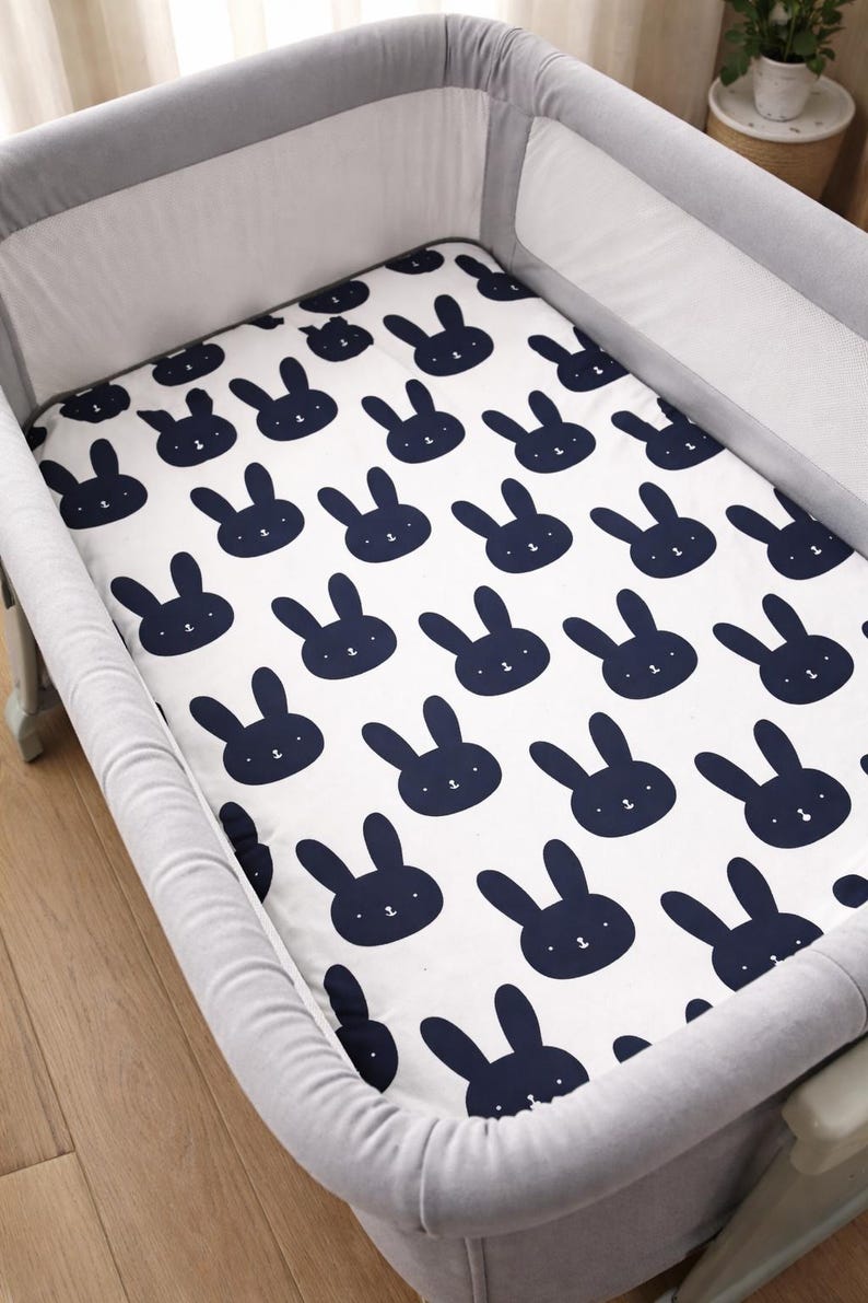 May include: A white cot sheet featuring a navy blue rabbit design. The sheet is placed inside a grey cot. The rabbit pattern is repeated across the sheet.