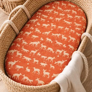 May include: Woven wicker baby bassinet with a deer-patterned orange mattress pad. The bassinet has rope handles and a white blanket draped over the side. The bassinet is designed for newborns and infants.