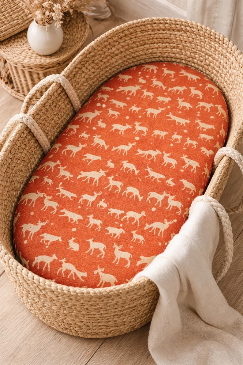 Woven wicker baby bassinet with a deer-patterned orange mattress liner. The bassinet has rope handles and a white blanket draped over the side. The bassinet is designed for newborns and infants.