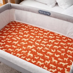 Puede incluir: Una cuna gris y blanca con una sábana bajera de color óxido. La sábana tiene un estampado de siluetas de ciervos color crema. La cuna tiene un panel lateral de malla y el texto "next to me" en la cabecera.