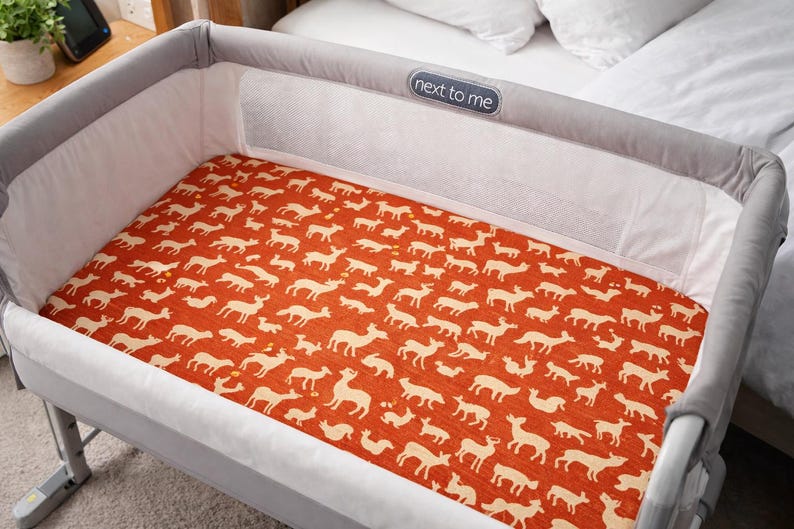 A grey and white baby bassinet with a rust-coloured fitted sheet featuring a repeating pattern of cream-coloured deer silhouettes. The bassinet has a mesh side panel and the text "next to me" on the headboard.