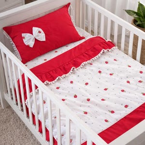 Puede incluir: Una cuna blanca con ropa de cama roja y blanca. La almohada roja tiene un lazo blanco con detalles de fresas. La manta blanca tiene un estampado de fresas y ribete rojo. La cuna está en una habitación con una cómoda blanca y una planta.