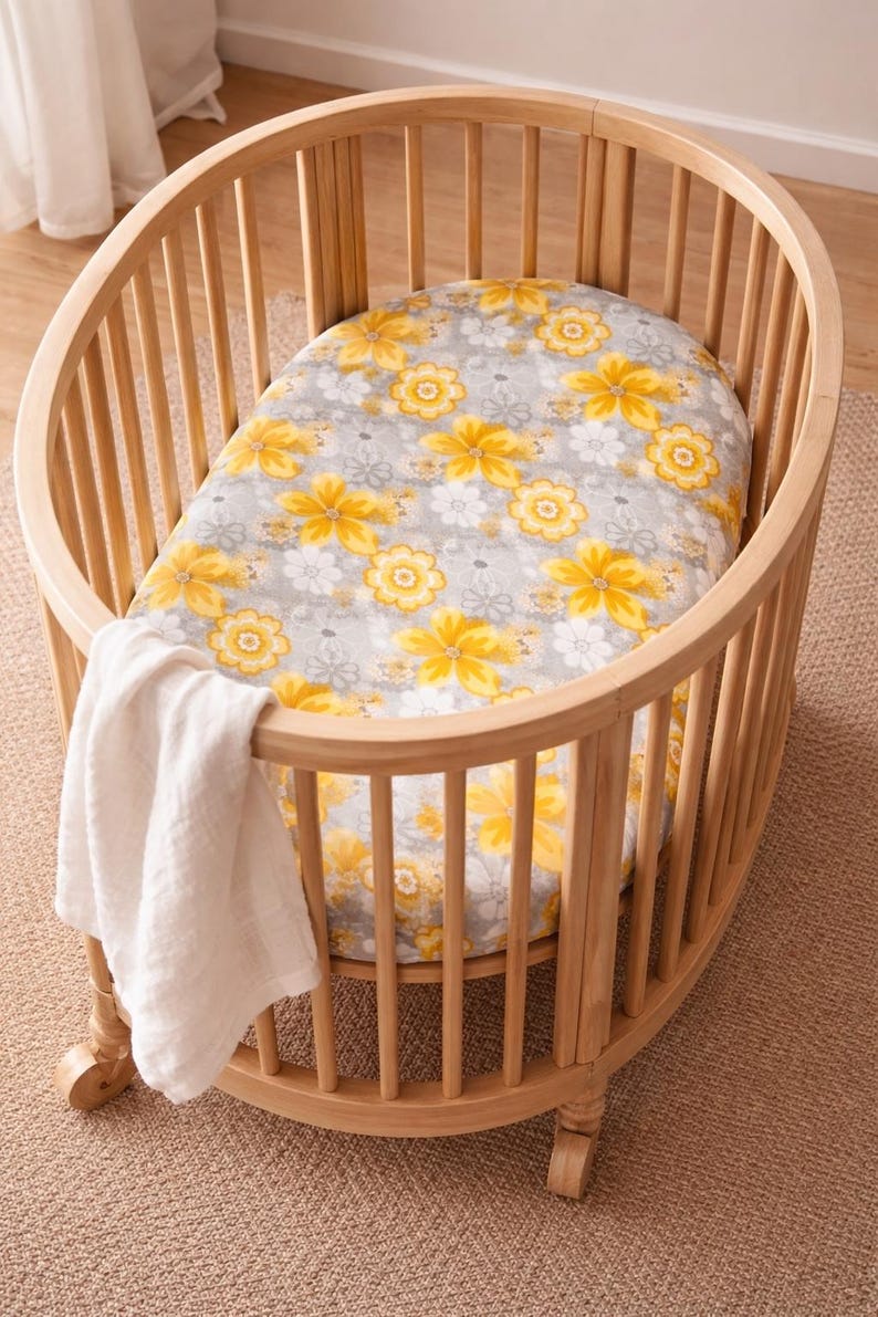 Puede incluir: Una cuna ovalada de madera clara con una s&aacute;bana estampada con flores amarillas y blancas sobre un fondo gris. Una manta blanca cuelga por un lado. La cuna est&aacute; sobre una alfombra marr&oacute;n claro.