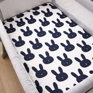 May include: A white cot sheet featuring a navy blue bunny face pattern. The sheet is placed inside a grey cot with a white interior.