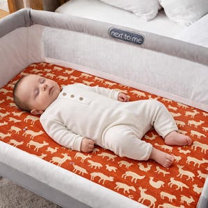 Puede incluir: Un beb&eacute; dormido con un body blanco yace en una cuna gris y blanca. La cuna tiene una s&aacute;bana roja estampada con siluetas de animales blancos. La cuna est&aacute; al lado de una cama con ropa de cama blanca.