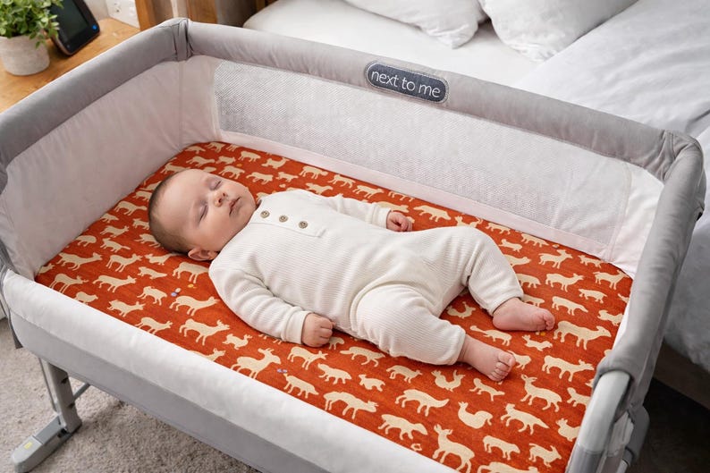 Puede incluir: Un beb&eacute; dormido con un body blanco yace en una cuna gris y blanca. La cuna tiene una s&aacute;bana roja estampada con siluetas de animales blancos. La cuna est&aacute; al lado de una cama con ropa de cama blanca.