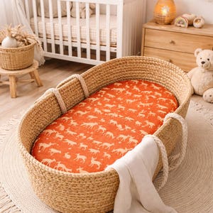 Puede incluir: Cuna de beb&eacute; de mimbre tejido con una s&aacute;bana bajera naranja con un estampado de ciervos blancos. La cuna tiene asas de cuerda y se apoya sobre una alfombra redonda con flecos. Una cuna blanca est&aacute; al fondo.