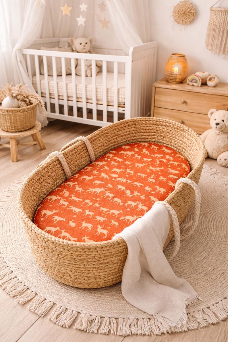 Puede incluir: Cuna de beb&eacute; de mimbre tejido con una s&aacute;bana bajera naranja con un estampado de ciervos blancos. La cuna tiene asas de cuerda y se apoya sobre una alfombra redonda con flecos. Una cuna blanca est&aacute; al fondo.