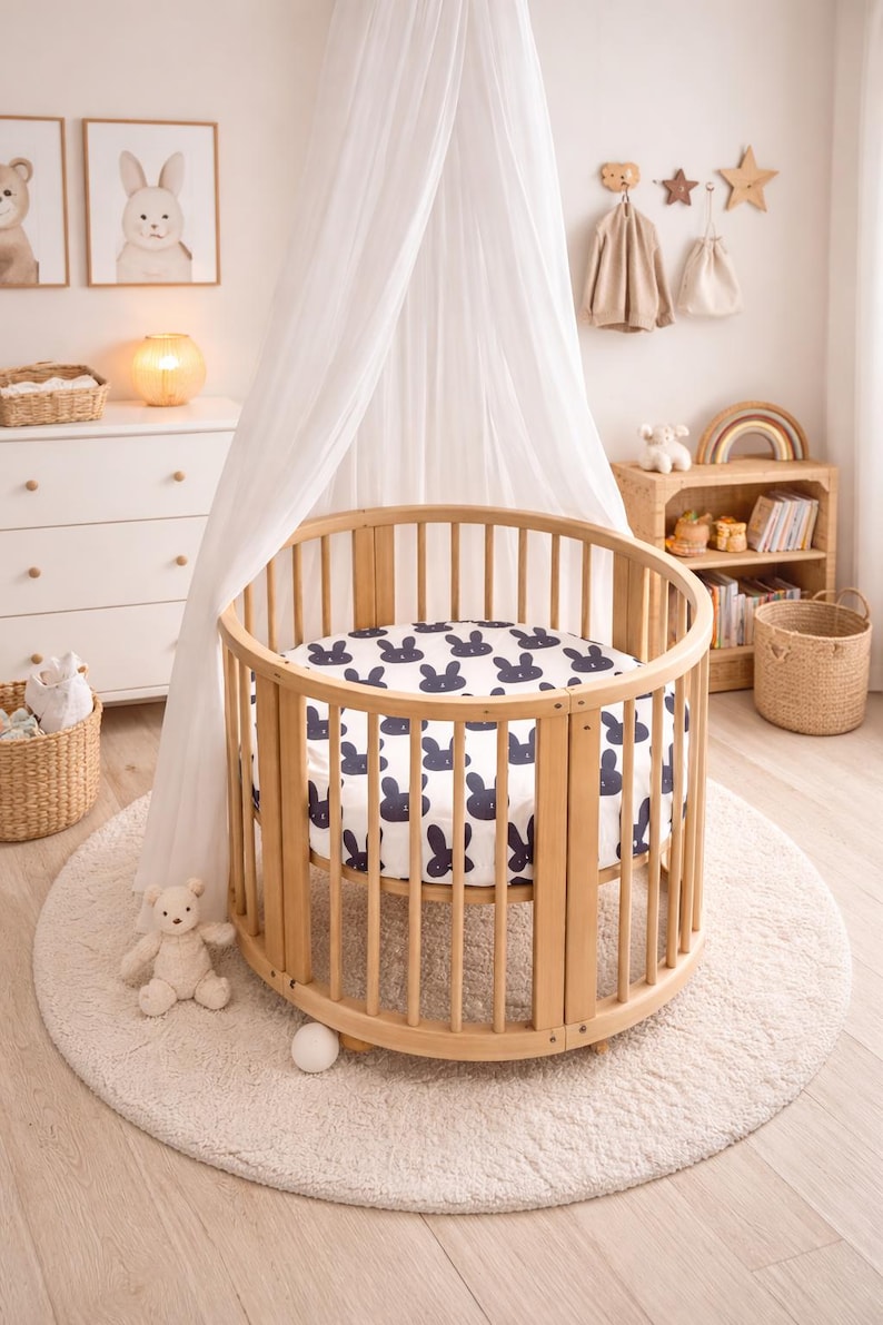 May include: A light wood-toned, circular baby cot with a navy blue bunny print sheet. The cot is on a cream rug, with a white canopy. The room is decorated with framed art and a teddy bear.