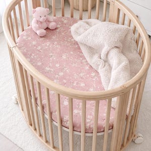 May include: A light wood oval baby crib with a pink floral patterned fitted sheet, a white fluffy blanket, and a pink teddy bear. The crib has vertical wooden slats and sits on small white wheels.