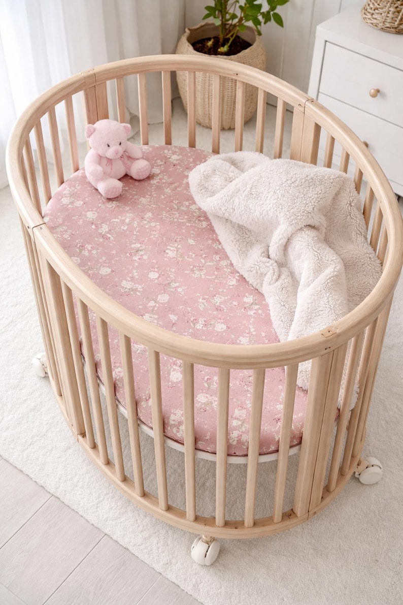 May include: A light wood oval baby crib with a pink floral patterned fitted sheet, a white fluffy blanket, and a pink teddy bear. The crib has vertical wooden slats and sits on small white wheels.