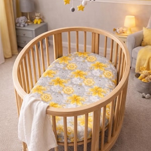 Puede incluir: Una cuna ovalada de madera con un colch&oacute;n con estampado floral y una manta blanca. La cuna est&aacute; en una guarder&iacute;a con una combinaci&oacute;n de colores amarillo y gris. Un m&oacute;vil con estrellas y nubes cuelga encima.