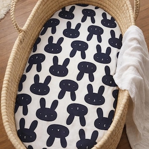 May include: A woven wicker bassinet with a white sheet featuring a navy blue bunny pattern. A white muslin blanket is draped over the side. The bassinet has two handles and is placed on a wooden floor.