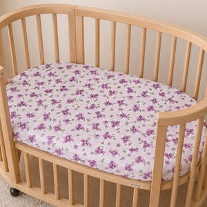 May include: A light wood oval crib with a floral patterned sheet. The sheet features a repeating pattern of purple flowers on a light pink background. The crib has vertical wooden slats and is on wheels.