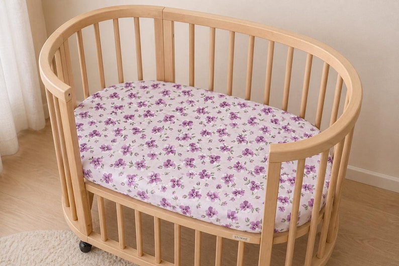 May include: A light wood oval crib with a floral patterned sheet. The sheet features a repeating pattern of purple flowers on a light pink background. The crib has vertical wooden slats and is on wheels.