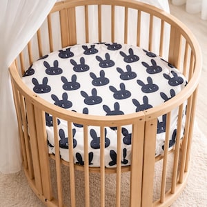 May include: A light wood-toned, oval baby cot with a white canopy. The cot features a fitted sheet with a navy blue bunny pattern. The cot is on a light-coloured rug.