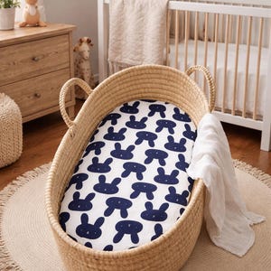 May include: A woven wicker baby bassinet with a navy blue bunny pattern sheet. The bassinet sits on a round jute rug with a fringed edge. A white muslin blanket is draped over the side.
