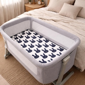 May include: A grey and white baby bassinet with a navy blue bunny pattern on the mattress. The bassinet is next to a bed with beige bedding. The room has a neutral colour palette.