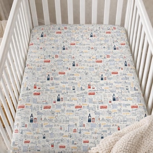 Puede incluir: Una cuna blanca con una sábana bajera con un estampado temático de Londres. La sábana tiene un diseño repetido de edificios, autobuses y monumentos en rojo, azul y amarillo sobre un fondo crema. Una manta color crema está sobre el lateral.