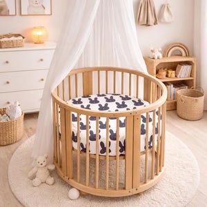 May include: A light wood-toned, circular baby cot with a navy blue bunny print sheet. The cot is on a cream rug, with a white canopy. The room is decorated with framed art and a teddy bear.