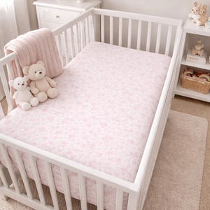 May include: A white crib with a pink floral patterned fitted sheet. A pink blanket and two teddy bears are on the side. The room has a white dresser, lamp, and artwork. The rug is a light beige color.