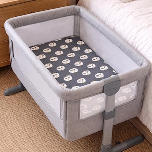 May include: A gray bassinet with a patterned mattress featuring white and yellow designs. The bassinet has a gray frame and is positioned next to a bed. The bassinet is supported by a gray stand.