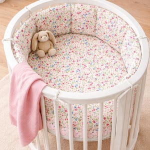 Op de afbeelding: Witte ovale babywieg met bloemmotief interieur en beddengoed. Een roze deken is over de zijkant gedrapeerd en een knuffelkonijn zit erin. De wieg is van hout.