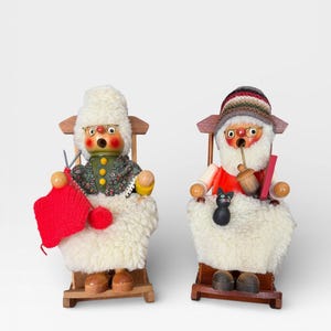 Puede incluir: Dos figuras de madera de personajes sentados, una tejiendo con lana roja y la otra fumando en pipa. Ambos llevan sombreros y están sentados en sillas de madera con cojines blancos y esponjosos. Las figuras tienen caras pintadas y visten ropa colorida.