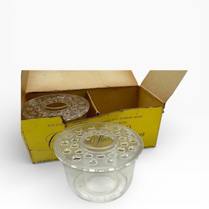 May include: Clear glass flower frog with multiple holes, designed to hold flower stems in place. The flower frog is sitting in front of a yellow cardboard box. The box has text that reads "For That..." and "Pink Flower".