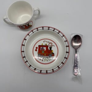 Puede incluir: Un juego de comedor para niños que presenta una taza blanca con asas, un cuenco pequeño y una cuchara. El cuenco y la taza tienen un diseño de tren rojo. El cuenco tiene un borde rojo y negro. La cuchara está en un envoltorio de plástico.
