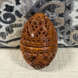 May include: A brown, egg-shaped wooden container featuring an intricate, carved pattern of interconnected circles and small, recessed holes. The object is composed of two halves that fit together, displaying a smooth, polished finish throughout.