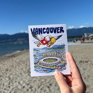 May include: A postcard featuring a cartoon seagull holding a golden trophy, with the word "Vancouver" above a stadium illustration. The postcard depicts a coastal city scene with mountains and boats, set against a blue sky and sandy beach.