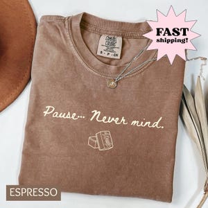 May include: An espresso-coloured short-sleeve t-shirt with a crew neckline. The shirt features the text 'Pause... Never mind.' in a light cream colour, accompanied by a small graphic of two mahjong tiles. A silver-toned necklace rests on the shirt.