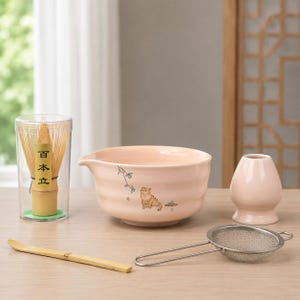 Puede incluir: Un juego de té matcha con un cuenco de cerámica rosa claro con una ilustración de gato, un batidor de bambú en un recipiente transparente, una cuchara de bambú, un jarrón pequeño y un colador de acero inoxidable. El juego es para preparar té matcha.