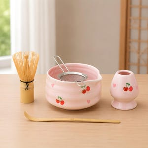 Op de afbeelding: Een matcha-theeset met een lichtroze keramische kom, garde, lepel en een kleine keramische houder. De kom en houder zijn versierd met kersen- en hartmotieven. De garde is gemaakt van bamboe.