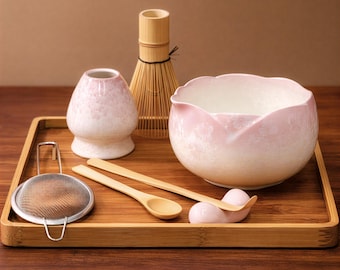May include: A matcha tea set on a bamboo tray. The set includes a pink and white floral bowl, a whisk, a whisk holder, a sifter, a matcha bowl, and two wooden spoons. The bowl has a floral design. The set is arranged on a bamboo tray.