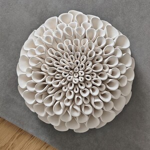 Può includere: Un'opera d'arte da parete in ceramica bianca a forma di fiore di dalia. Il fiore ha più strati di petali, creando un effetto strutturato e tridimensionale. L'opera è ambientata su uno sfondo grigio.