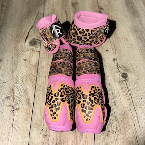 May include: A set of four horse leg boots and bell boots featuring a pink neoprene base with a brown and black leopard print pattern. The boots are accented with rows of small, shimmering crystals and a circular black patch with a white letter R.