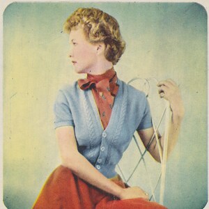 May include: A woman in a light blue cardigan with short sleeves and a red scarf with a floral pattern. She is wearing a red skirt and sitting in a white chair.