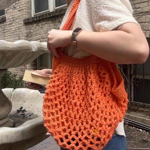 May include: An orange crochet tote bag with a shoulder strap. The bag has a net-like design and is held by a person. The bag is made of a thick yarn and has a simple, open weave. The bag is a bright, solid orange color.