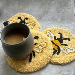 May include: A gray mug filled with a light brown drink sits on a yellow, round coaster. The coaster has a tufted design with a black and yellow bee illustration. Two more bee coasters are visible.