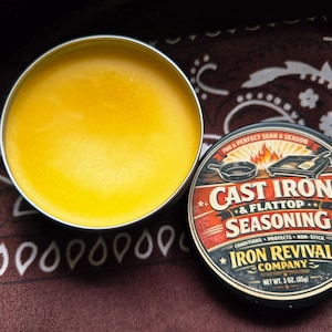 May include: A round metal tin filled with yellow seasoning next to a tin with a label that reads "Cast Iron & Flattop Seasoning". The label has a red, white, and black design with the text "Iron Revival Company".