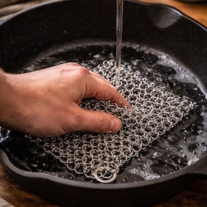 May include: A stainless steel chainmail scrubber is used to clean a black cast iron skillet. Water streams from above, creating suds. The scrubber is held in place by a hand. The skillet sits on a wooden surface.