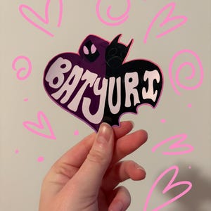May include: A heart-shaped sticker featuring a split design with a purple hooded figure on the left and a black bat-like silhouette on the right. The text BATYURI is written in large white letters across the center. The sticker is surrounded by pink hand-drawn hearts and swirls.