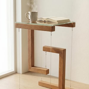 Könnte beinhalten: Einzigartiger Beistelltisch aus Holz mit Glasplatte und geometrischem Design. Eine Tasse Kaffee und ein offenes Buch liegen darauf. Der Tisch scheint an dünnen Drähten zu schweben. Das Holz hat einen warmen, natürlichen Farbton. Der Tisch ist ca. 76 cm hoch.
