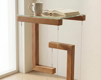 May include: A unique wooden side table with a glass top, featuring a geometric design. A cup of coffee and an open book rest on the table. The table is suspended by thin wires, creating a floating illusion. The wood has a warm, natural tone.