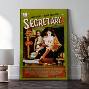 May include: A framed movie poster for "Secretary" with a green border. The poster features James Spader and Maggie Gyllenhaal. The title is in large white letters with red hearts. The poster has a vintage aesthetic.