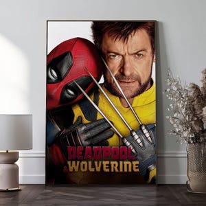 K&ouml;nnte beinhalten: Ein gerahmtes Poster mit Deadpool und Wolverine. Deadpool tr&auml;gt seine rote Maske und seinen Anzug, w&auml;hrend Wolverine in einem gelben und blauen Anzug mit seinen Krallen zu sehen ist. Das Poster tr&auml;gt die Worte "Deadpool & Wolverine" in Rot.