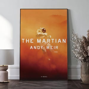 May include: A framed poster featuring the book cover of "The Martian" by Andy Weir. The artwork depicts an astronaut in a white spacesuit against an orange and red background. The title and author's name are in white, with "A Novel" at the bottom.