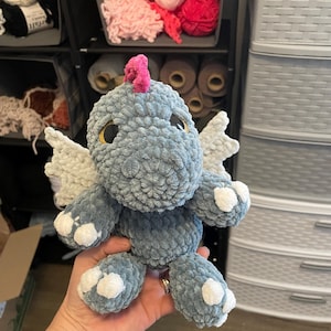 May include: A handmade, plush dragon toy in shades of gray, with white wings and accents. The dragon has large, round eyes and a pink crest. The toy is made of soft, textured yarn, and is held in a person's hand.